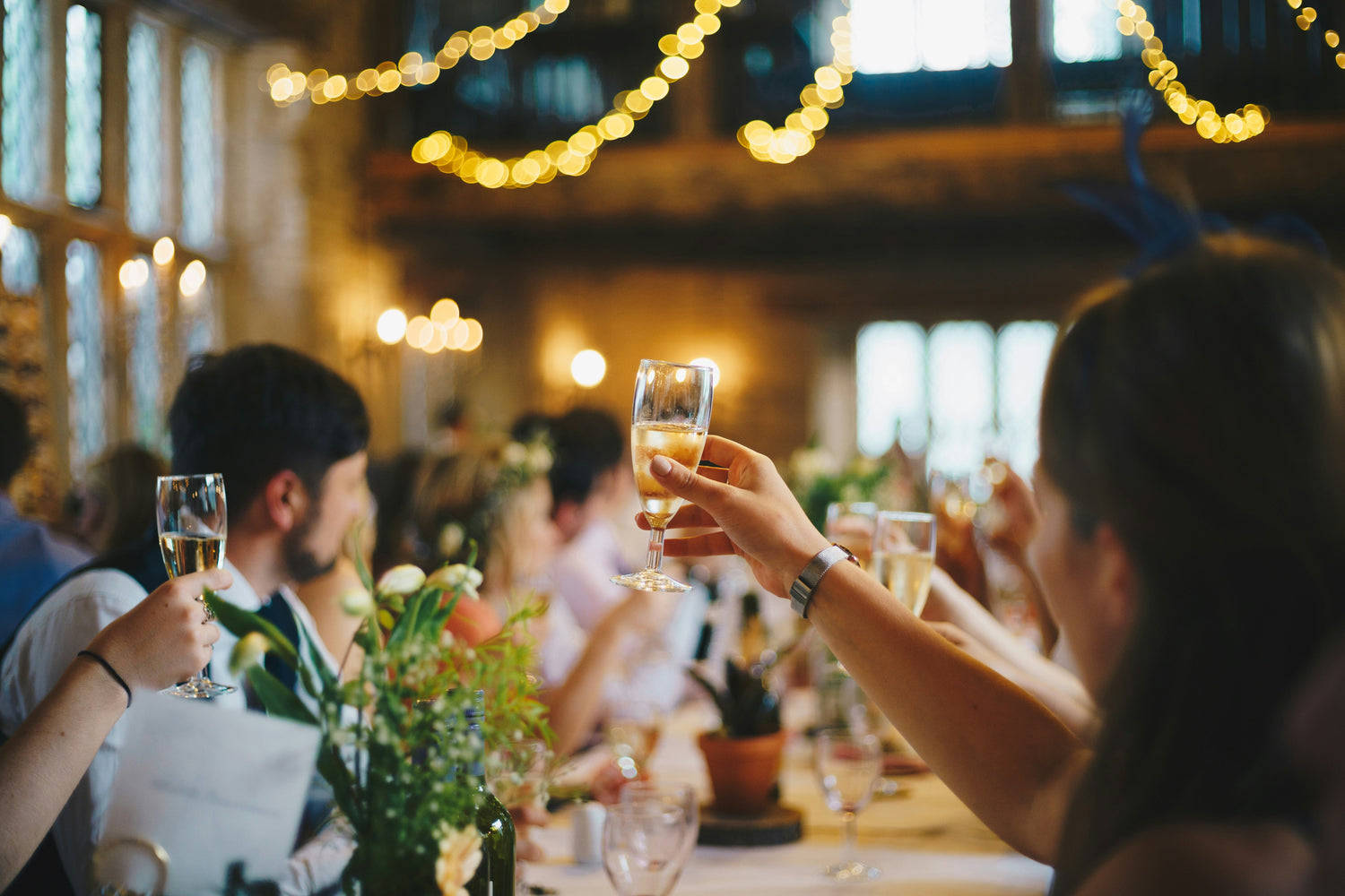 image shows people raising champagne flutes in a toast at a wedding. There are flowers on the tables and fairy lights in the background. 