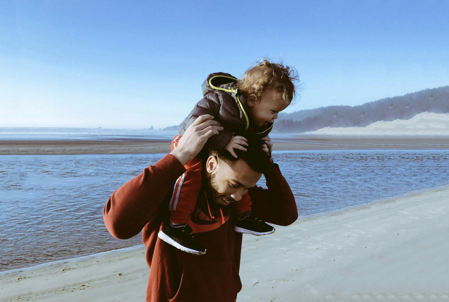 image shows a father on a beach with blue sea and blue sky in the background, he has his young son on his shoulders